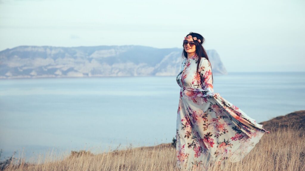 Femme portant une robe longue avec des motifs floraux roses, se tenant devant un paysage côtier avec un bandeau de fleurs et des lunettes de soleil.