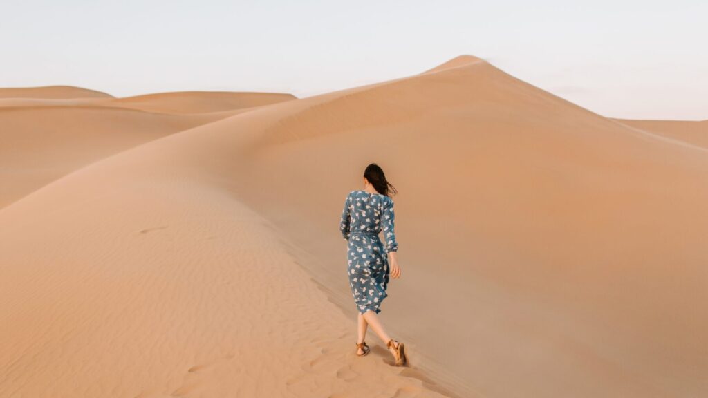 Femme de dos, vêtue d'une robe fleurie bleue, marchant à travers les dunes de sable du désert, les cheveux soufflés par le vent.