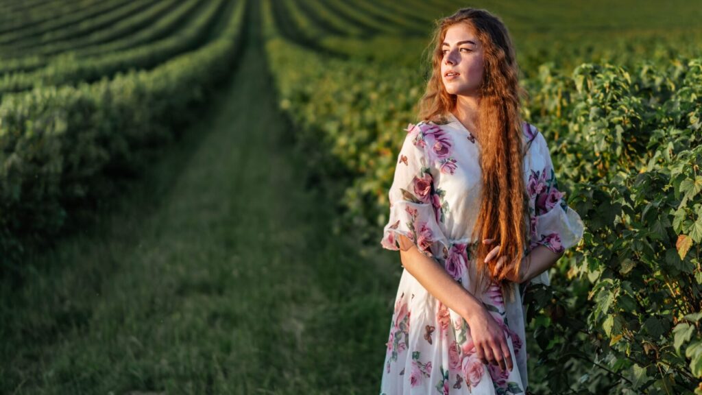 Jeune femme avec de longs cheveux roux, portant une robe fleurie blanche avec des motifs roses, posant dans un champ de vignes verdoyantes.