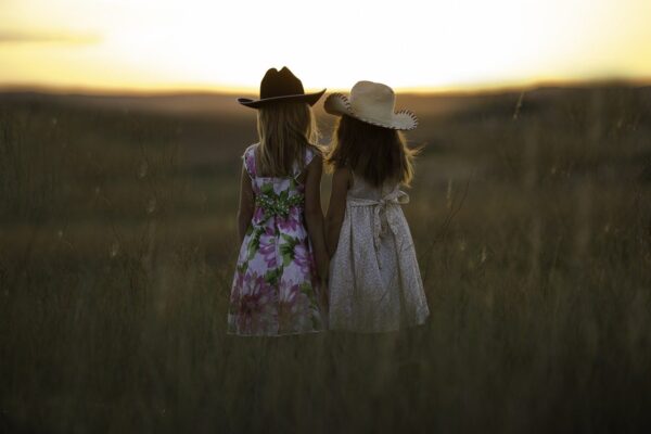 Deux petites filles portant des robes fleuries et des chapeaux de cowboy regardent un coucher de soleil dans un champ.