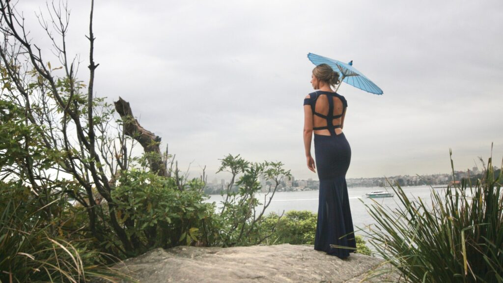 Femme en robe dos nu bleue, debout sur un rocher avec un parapluie, face à un paysage urbain et marin.